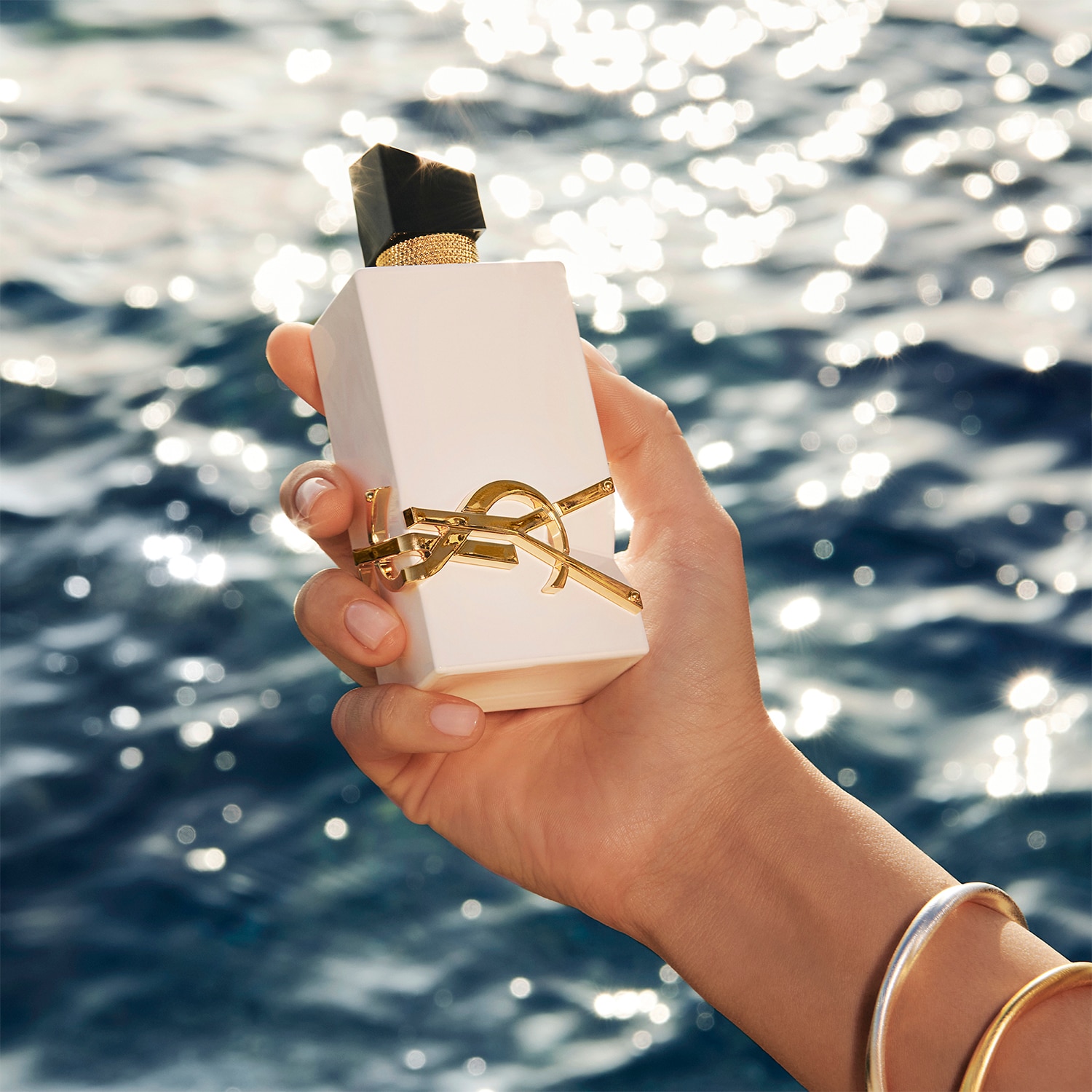Mano femenina sosteniendo el frasco blanco y dorado de la nueva fragancia Libre sin alcohol con el reflejo del sol sobre el agua de fondo.